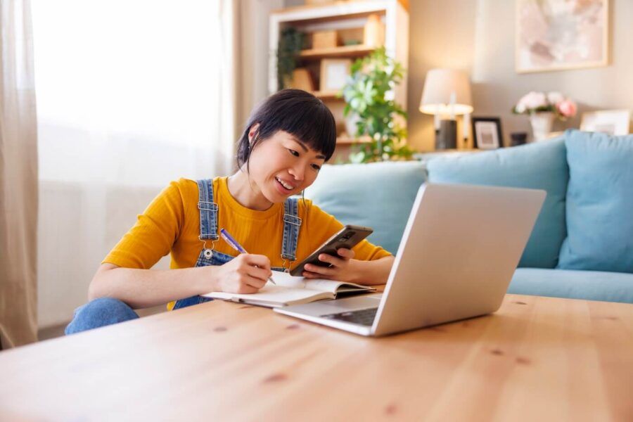 Smiling woman is using her laptop on a coffee table while holding her smartphone and making notes