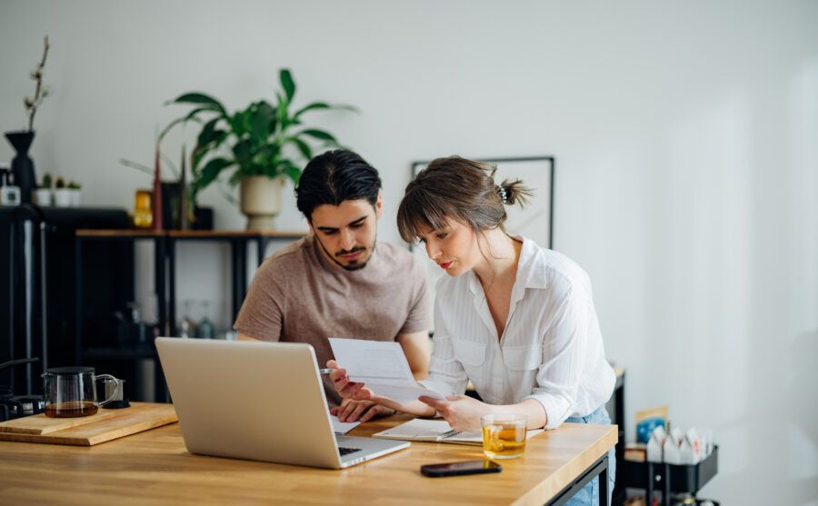 Focused couple sitting at a kitchen table, intently reviewing their finances together. They are using a laptop, surrounded by documents, a calculator, and mugs