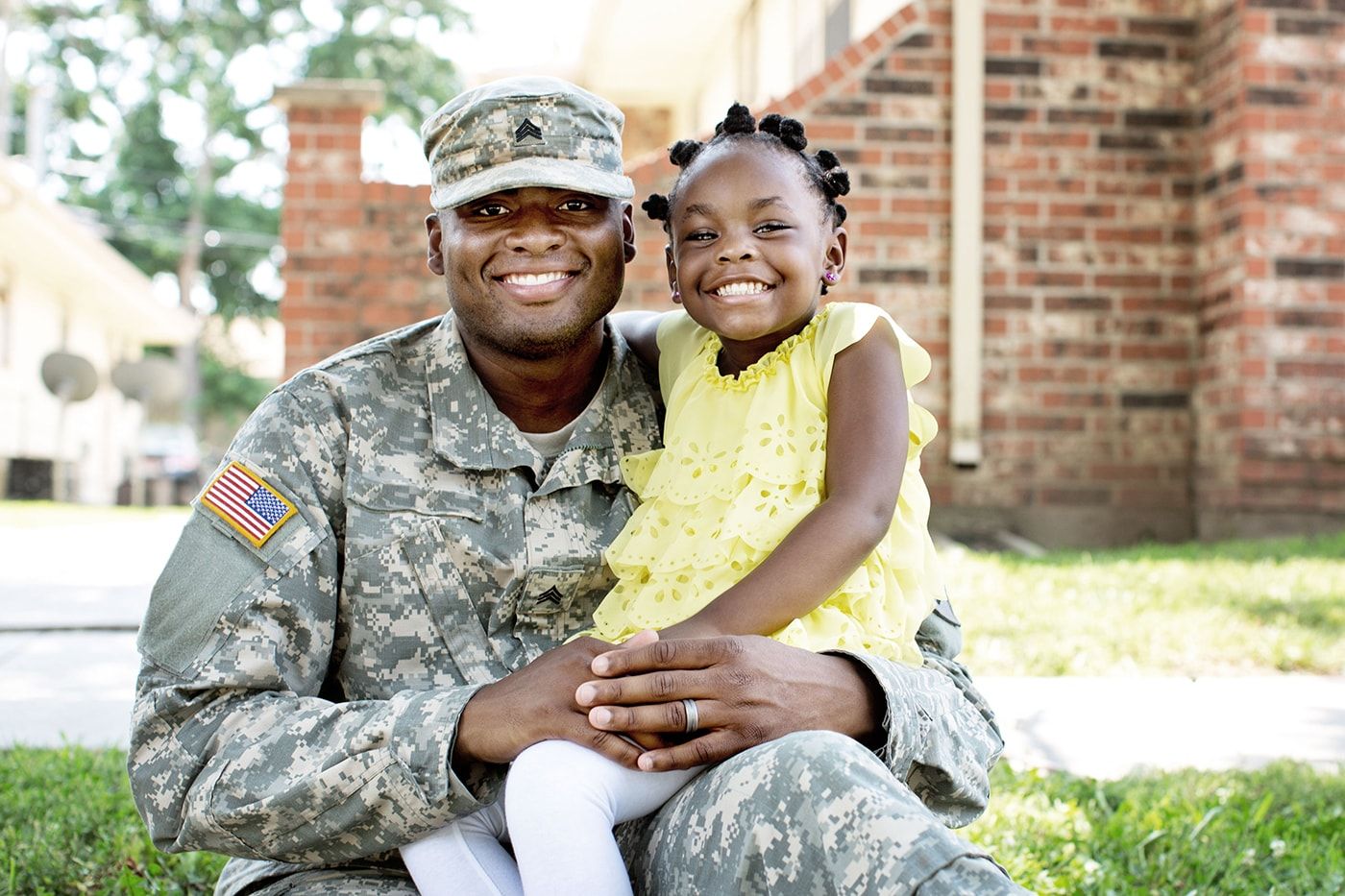 A military father smiles while his daughter wearing a yellow dress sits on his lap outside.