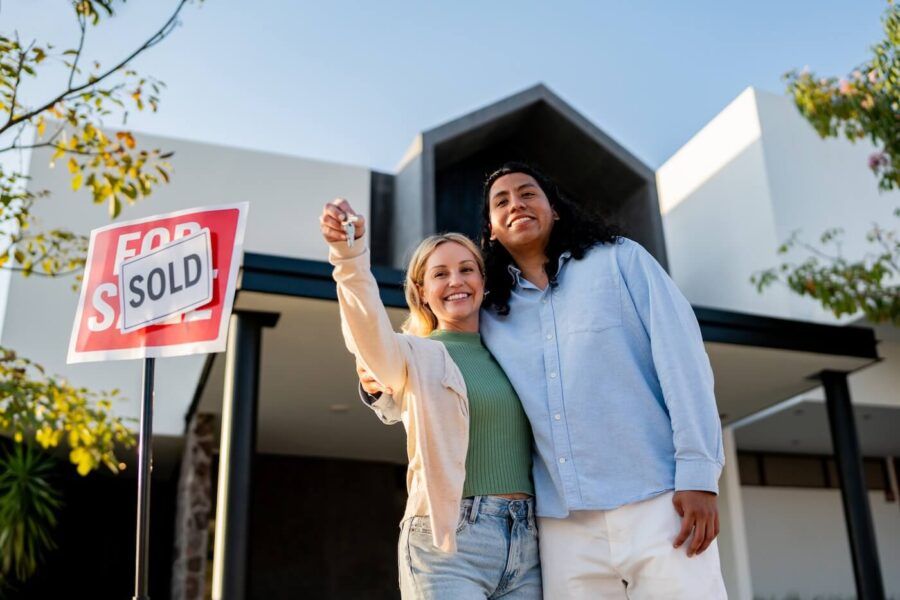Happy couple holding the keys from their new house while standing next to the 'Sold' sign