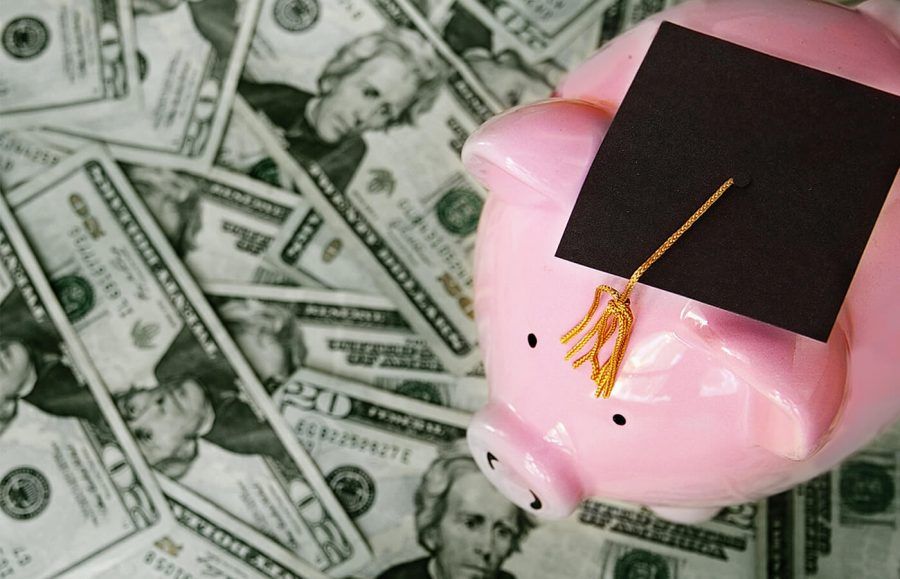 A piggy bank wearing a graduation cap sitting on a pile of American money.