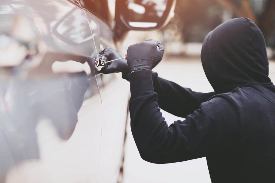 A burglar lock picks a car door while wearing a black jacket and gloves.