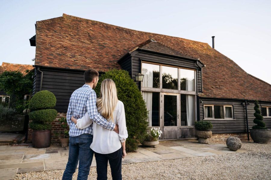 A couple embrace each other as they look at a home that has black walls and a brown roof.