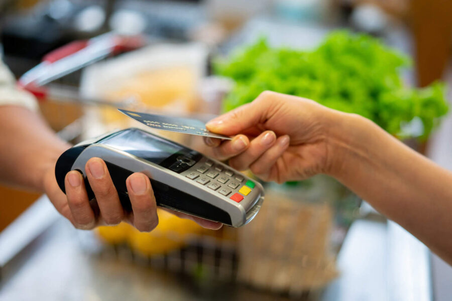 A customer holds up their credit card to tap on the card reader while their groceries are in a basket in the background.