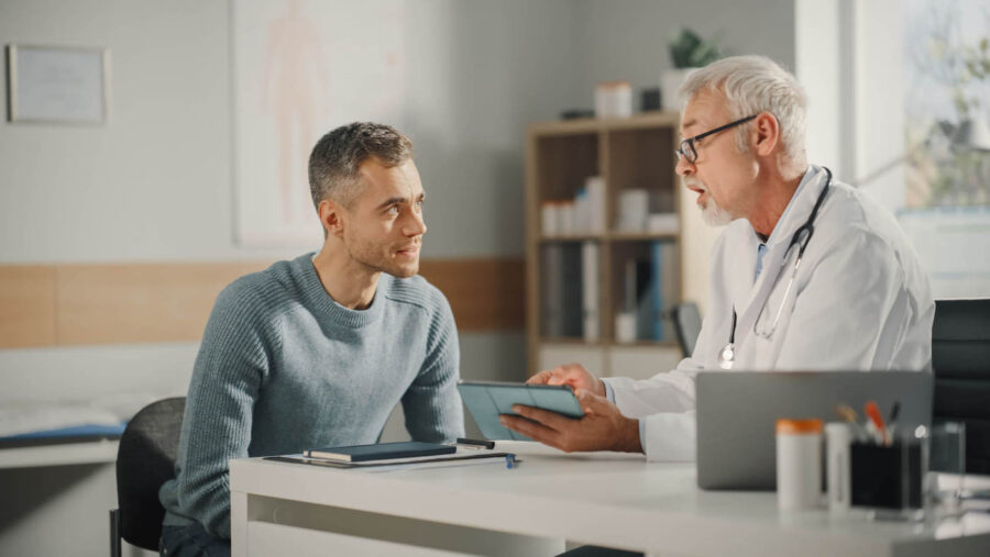 A doctor wearing a white coat is talking with a male patient at the doctor's office.