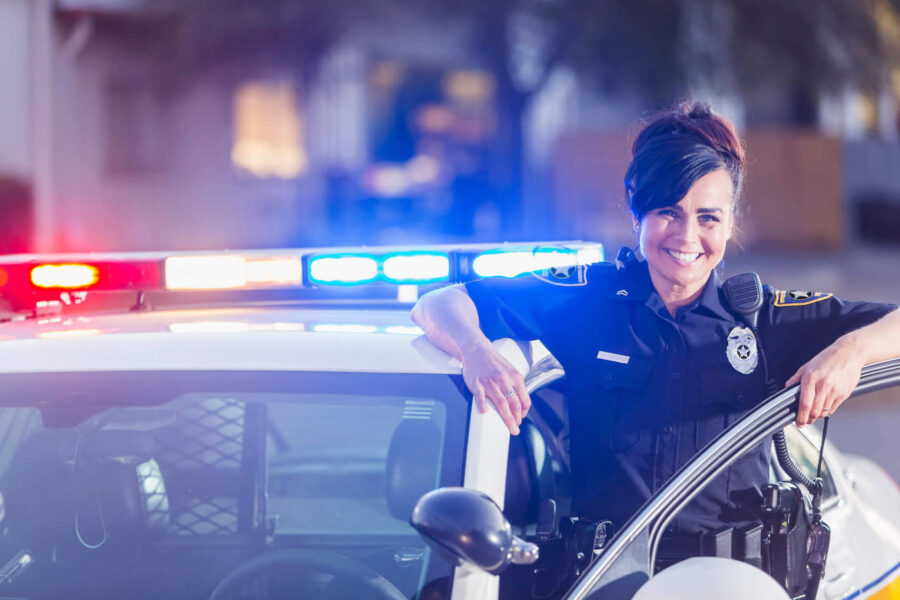 A female police officer wearing her uniform smiles while standing next to her police car.