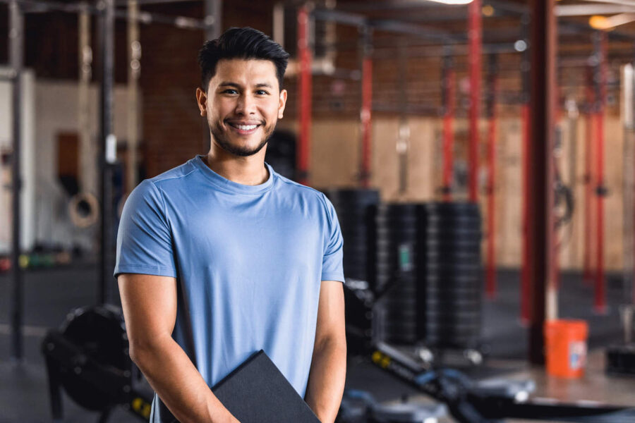 A fitness trainer wearing a blue shirt holds a folder and smiles at the gym.