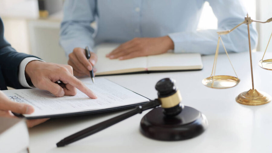 A hand points at a section of a contract for another person to sign with a gavel on the table.
