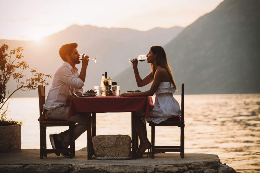 A man and a women are drinking wine and having dinner with the sunset and lake in the background.