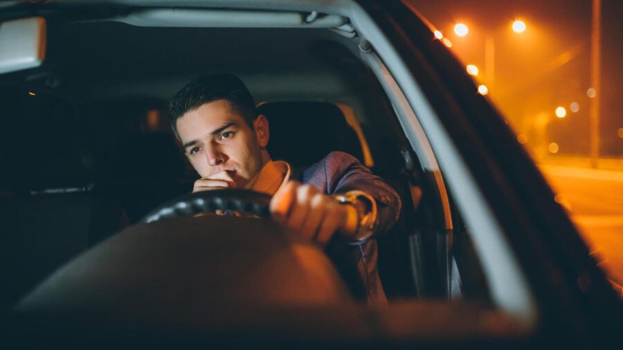 A man is driving at night with one hand on the wheel and his other hand on his mouth.