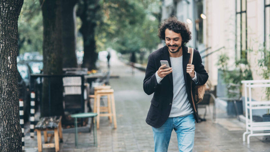 A man is walking outside while wearing his backpack as he looks at his phone and smiles.