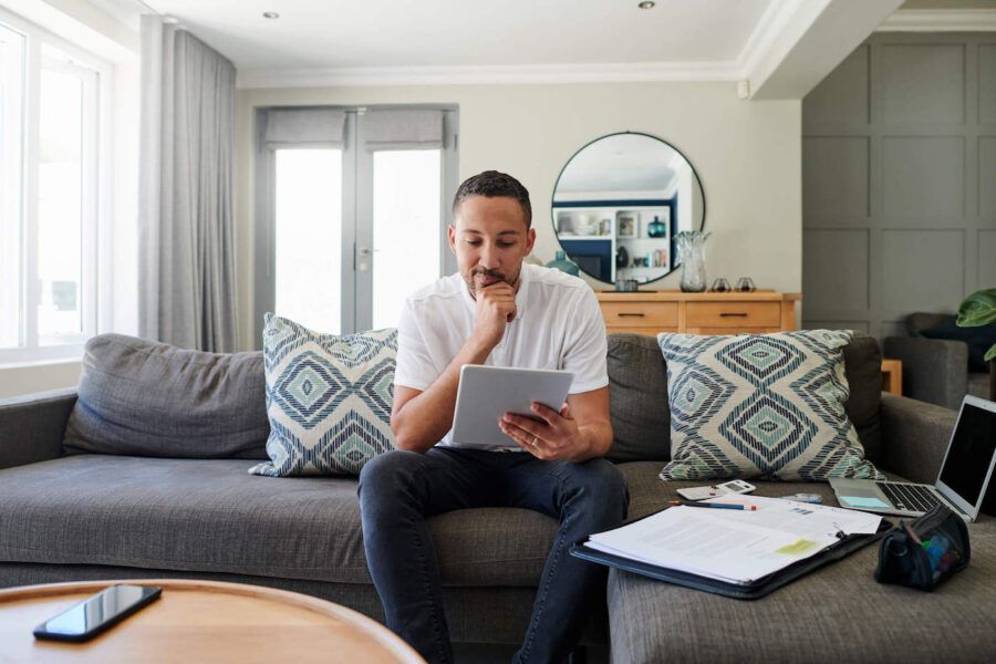 A man sitting on the couch looks at his tablet while a folder full of documents are off to the side.