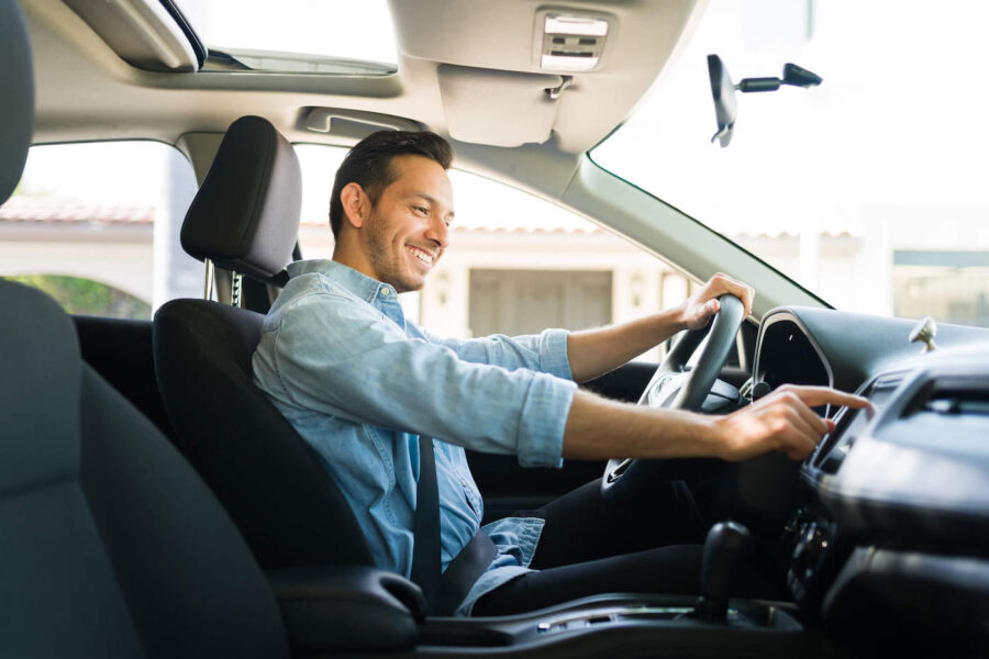 A man wearing a blue shirt smiles as he touches his radio while behind the wheel of his car.