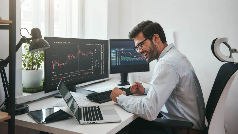 A man smiles while looking at stock charts on his computer from his home office.