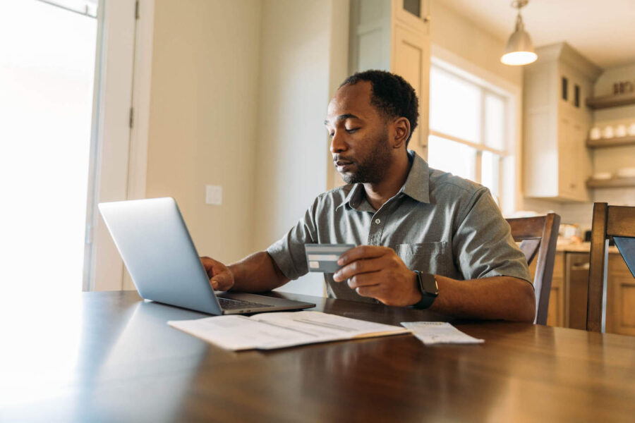 A man wearing a gray button-up shirt uses his laptop while holding his credit card at home.