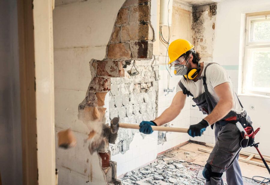 A man wearing a hard hat and safety glasses uses a sledgehammer to break down a wall of a home.