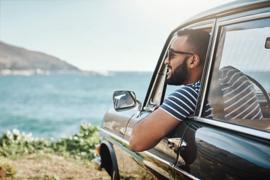 A man wearing sunglasses smiles as he looks towards the ocean from his car window.