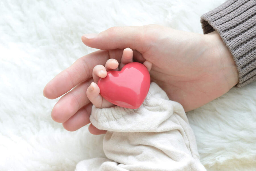 A parent has their hand under their child's hand that is holding a red heart.