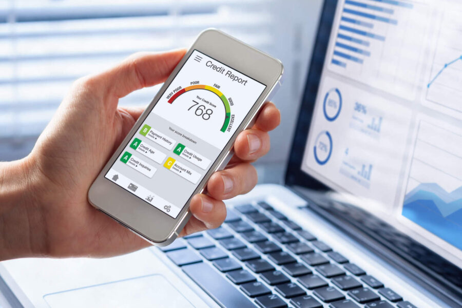 A person holds their phone that shows their current credit score while their laptop in the background shows data graphs.