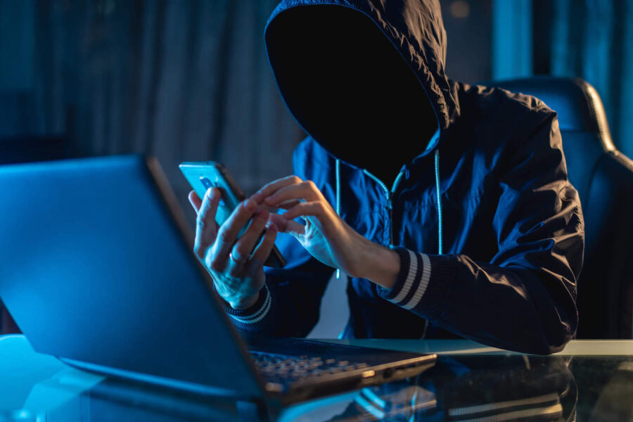 A person wearing a black hoodie is using a phone and laptop while their face is hidden.