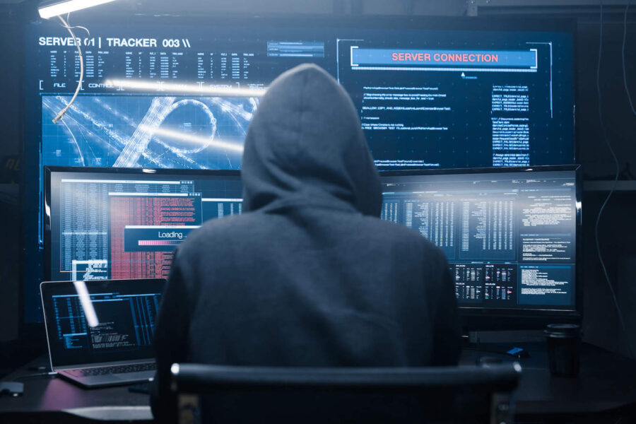 A person wearing a gray hoodie looks at multiple computer and tv displays showing dark web websites.