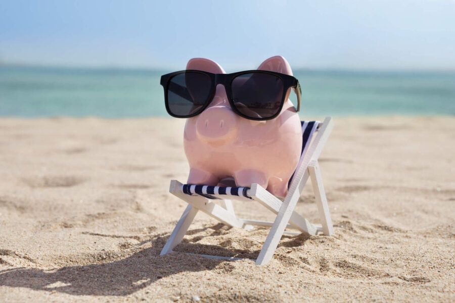 A pink piggy bank is sitting in a miniature beach chair while wearing sunglasses at the beach.