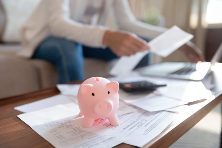 A pink piggy bank sits on top of documents on the living room table while someone type on a laptop in the background.
