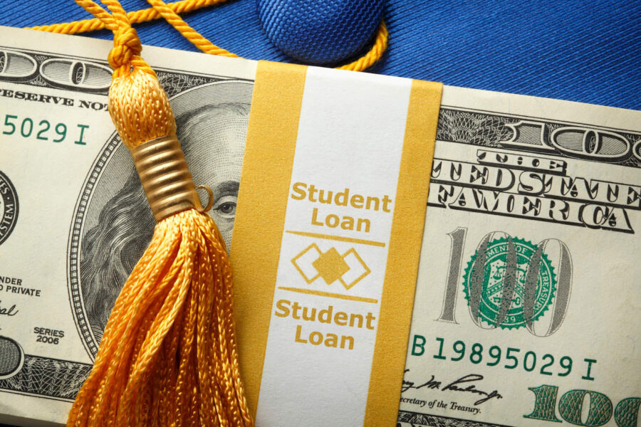 A stack of student loan cash sits on top of a blue graduation cap.
