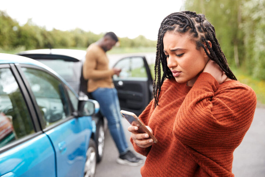 A woman wearing an orange sweater is holding her neck and using her phone with a car accident in the background.