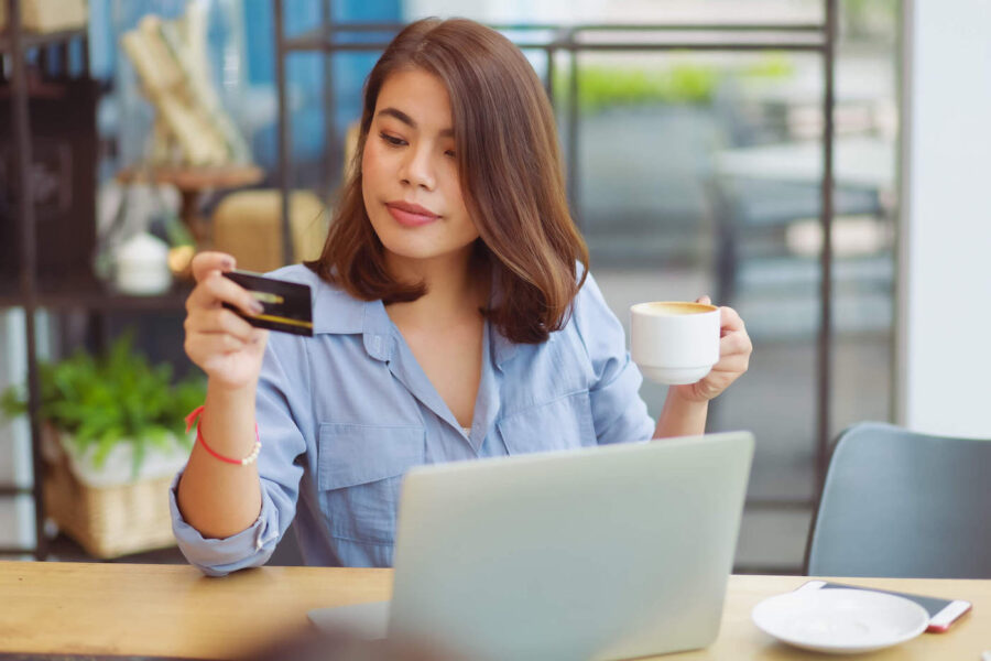 A woman holds a cup of coffee while looking at her credit card with her laptop on the table.