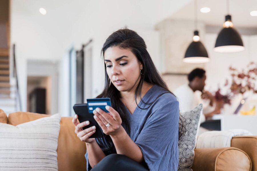 A woman sitting on the couch uses her phone while holding her credit card in her other hand.