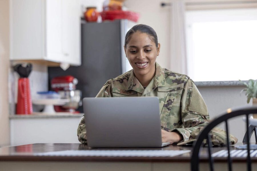 A woman in military uniform smiles as she uses her laptop.