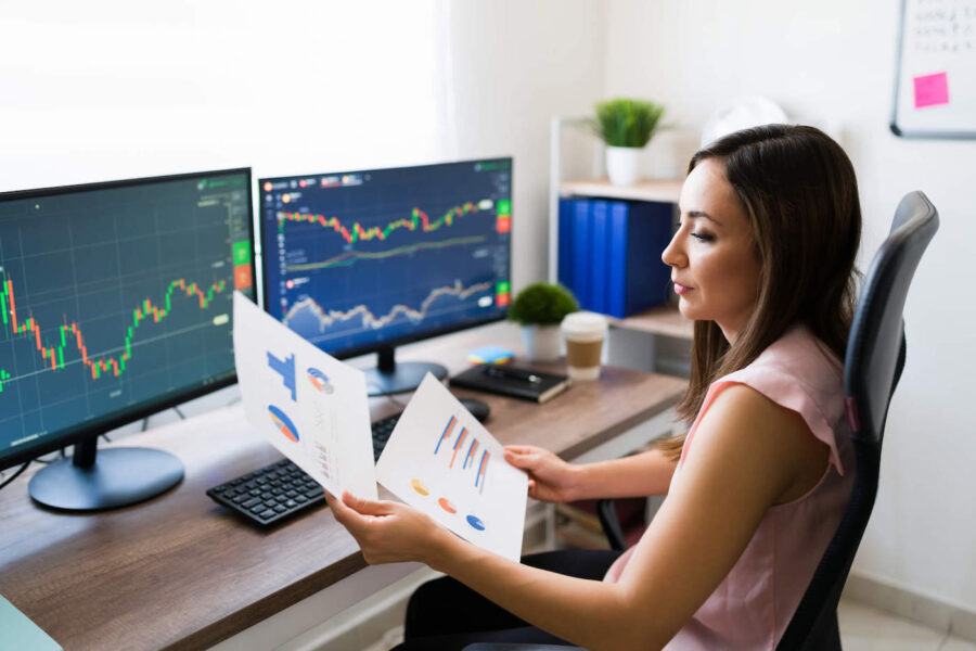 A woman is sitting at her desk looking at data documents while her desktop screens show stock charts.
