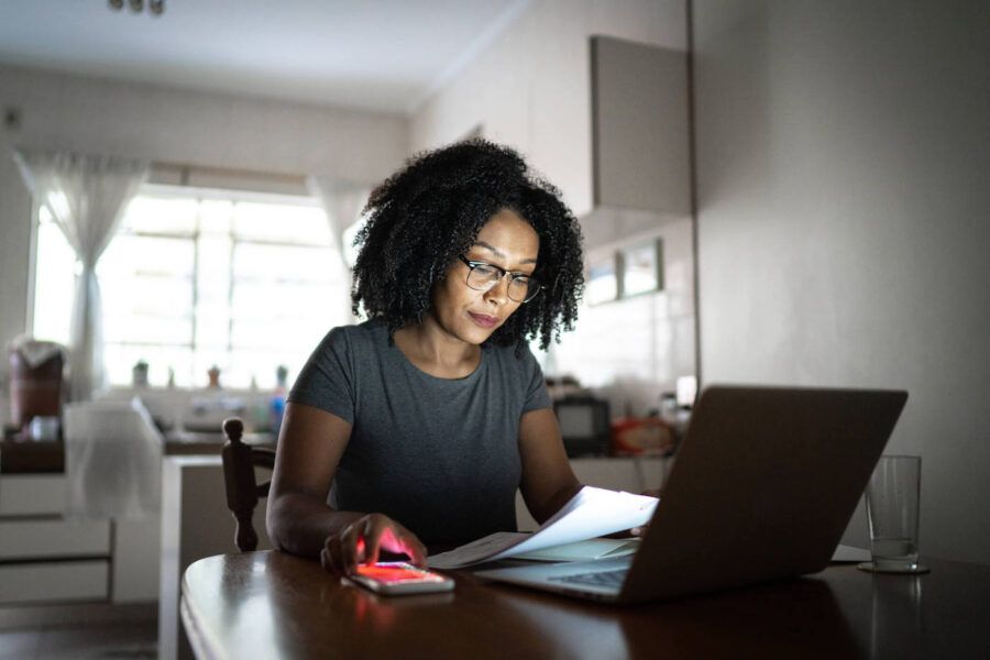 A woman wearing glasses looks at documents while her phone and laptop are on the kitchen table.