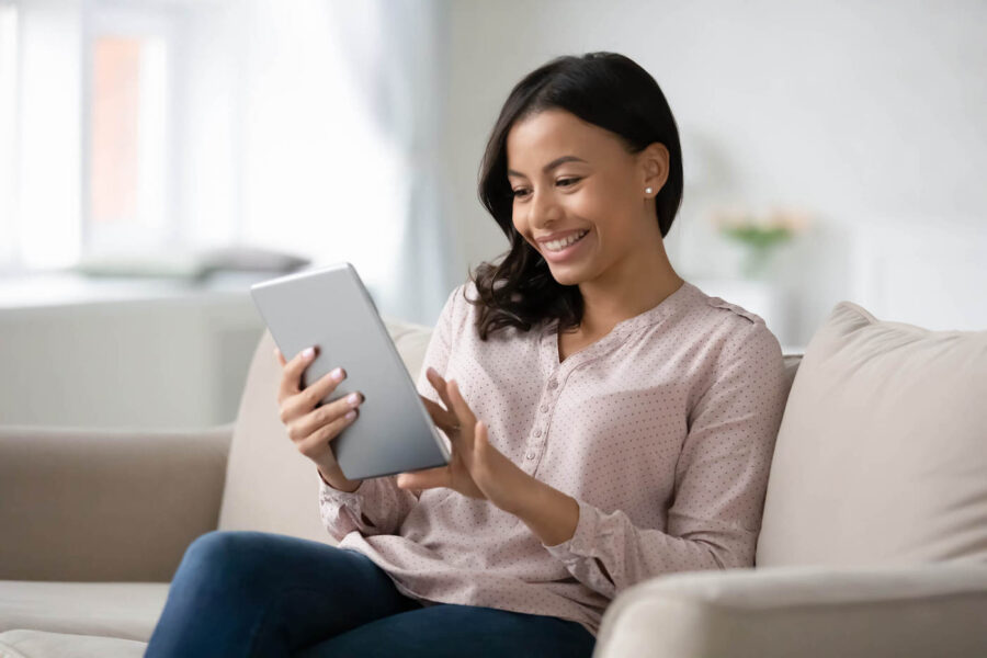 A woman sits at the couch while smiling and using her tablet.