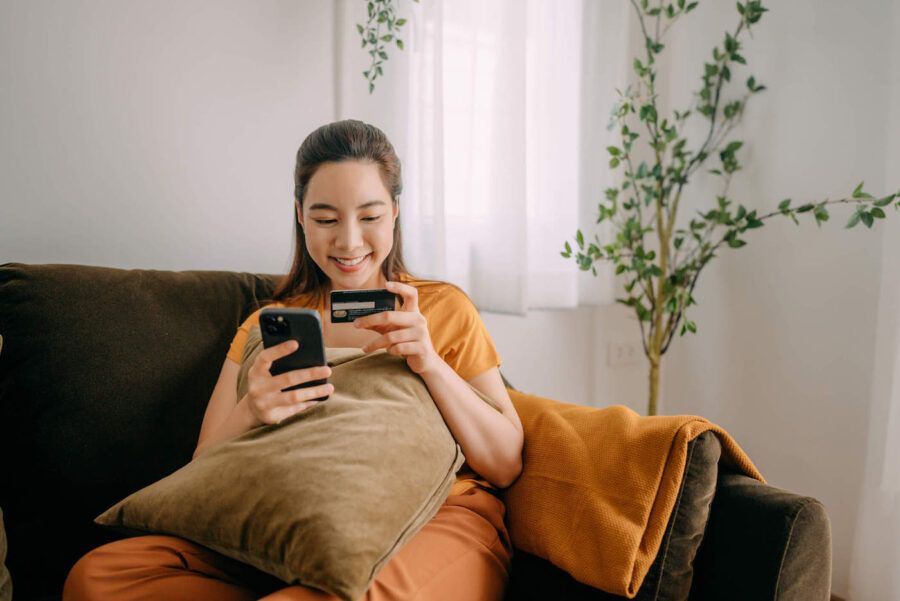 A woman sitting on the couch smiles at her credit card while also holding her phone.