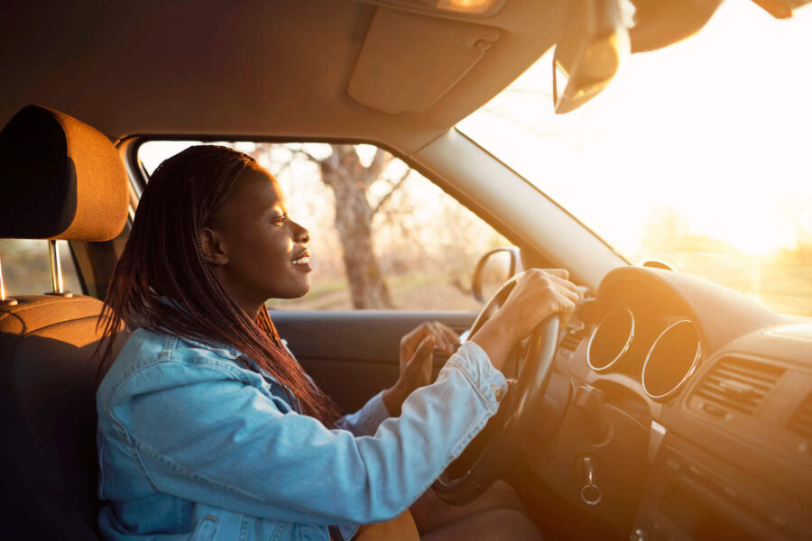 A woman wearing a blue denim jacket smiles while driving her car with the sunset in the background.