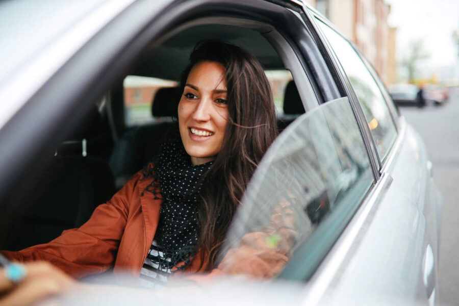 A woman wearing an orange jacket and black scarf smiles as she drives with her window down.