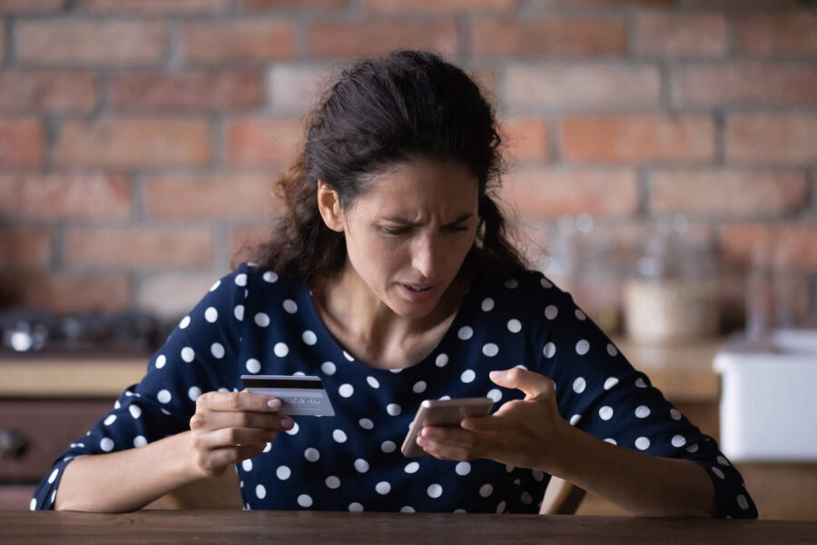 A woman wearing a polkadot shirt frowns while looking at her phone and holding her credit card.
