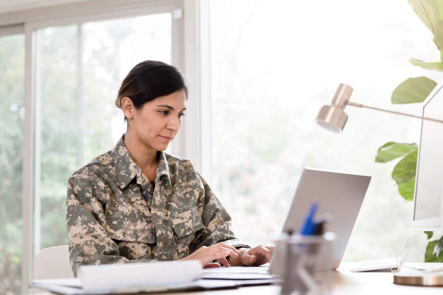 A woman wearing military uniform sits down at her desk and uses her laptop.