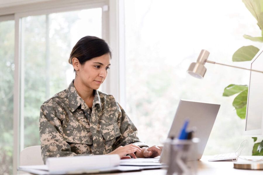 A woman wearing military uniform sits down at her desk and uses her laptop.