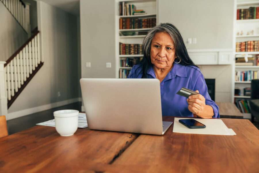 A woman wearing a purple shirt looks at her laptop while holding her credit card.