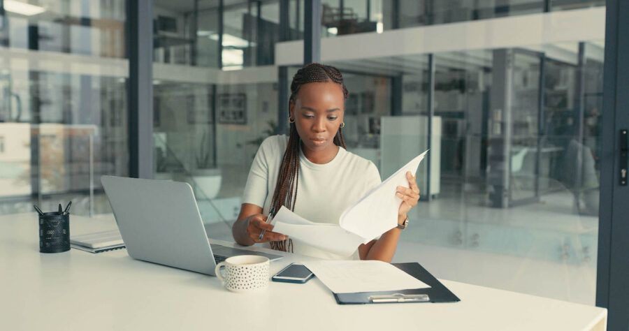 A woman with braids looks at documents while at the office with her laptop open at her desk.