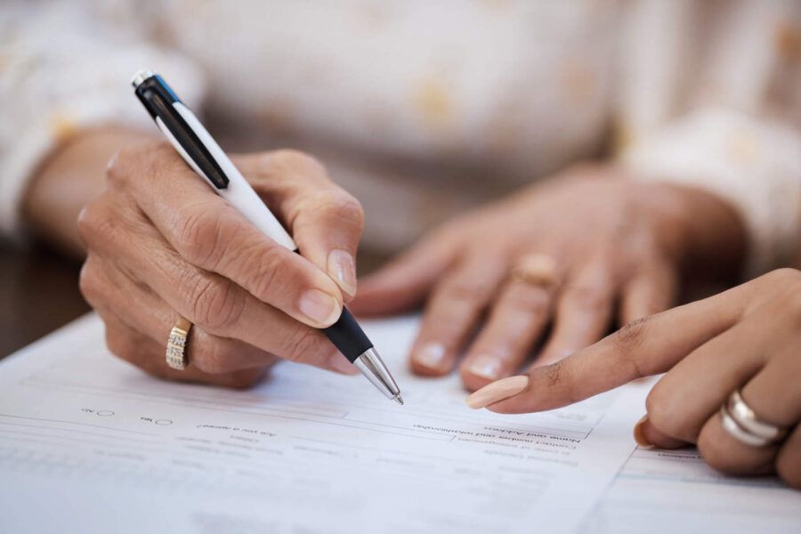A women wearing a ring is holding a pen to sign a document while another women points where to sign.