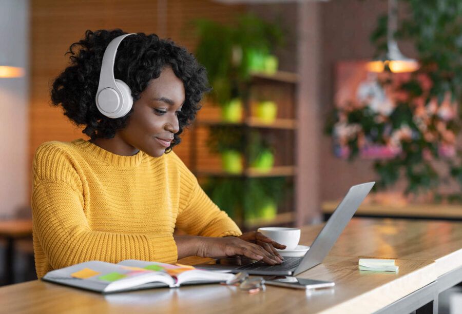 A women wearing a yellow sweater and white headphones is typing on her laptop with plants behind her.
