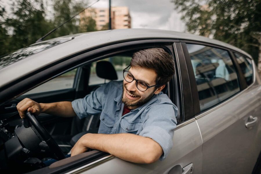 A young man wearing glasses smiles while looking outside of his car window.