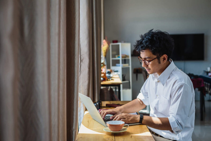 A young man wearing a white shirt and glasses uses his laptop at home.