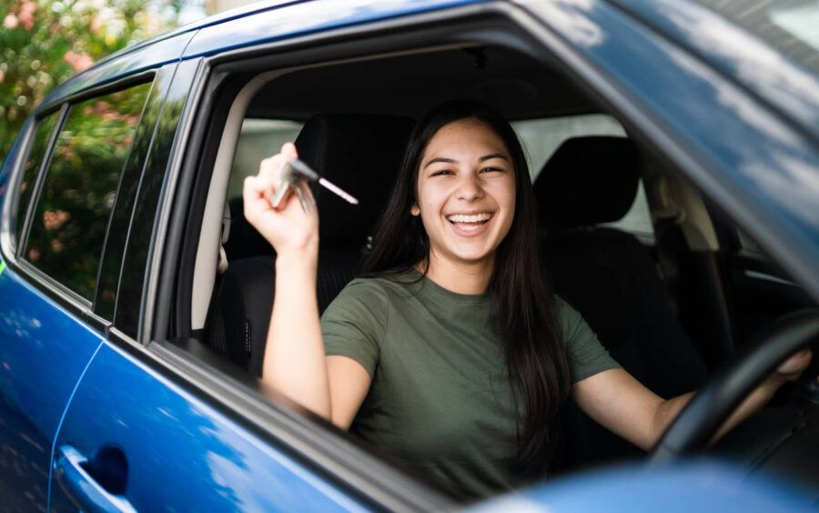 A young woman happily hold up her car keys while inside of her blue car.
