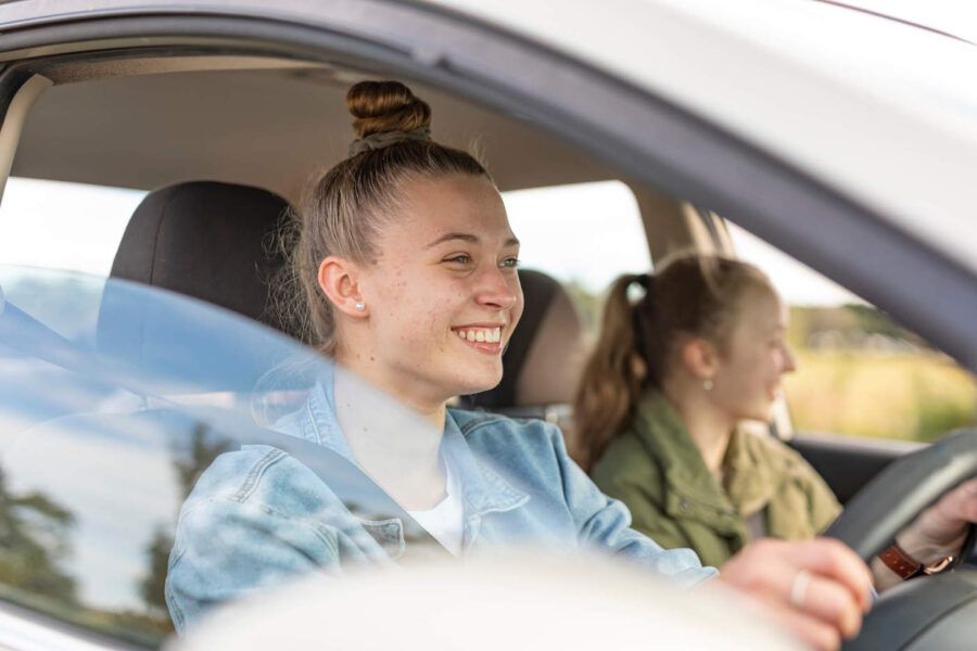 A young woman with blonde hair is smiling while driving with another young woman in the passenger seat.