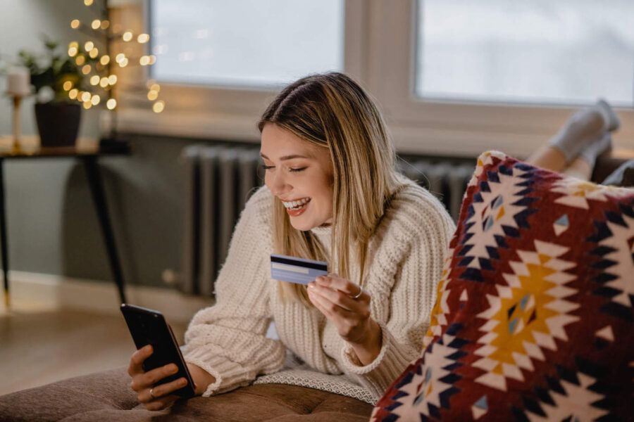A young woman wearing a white sweater laughs at her phone while holding her credit card.